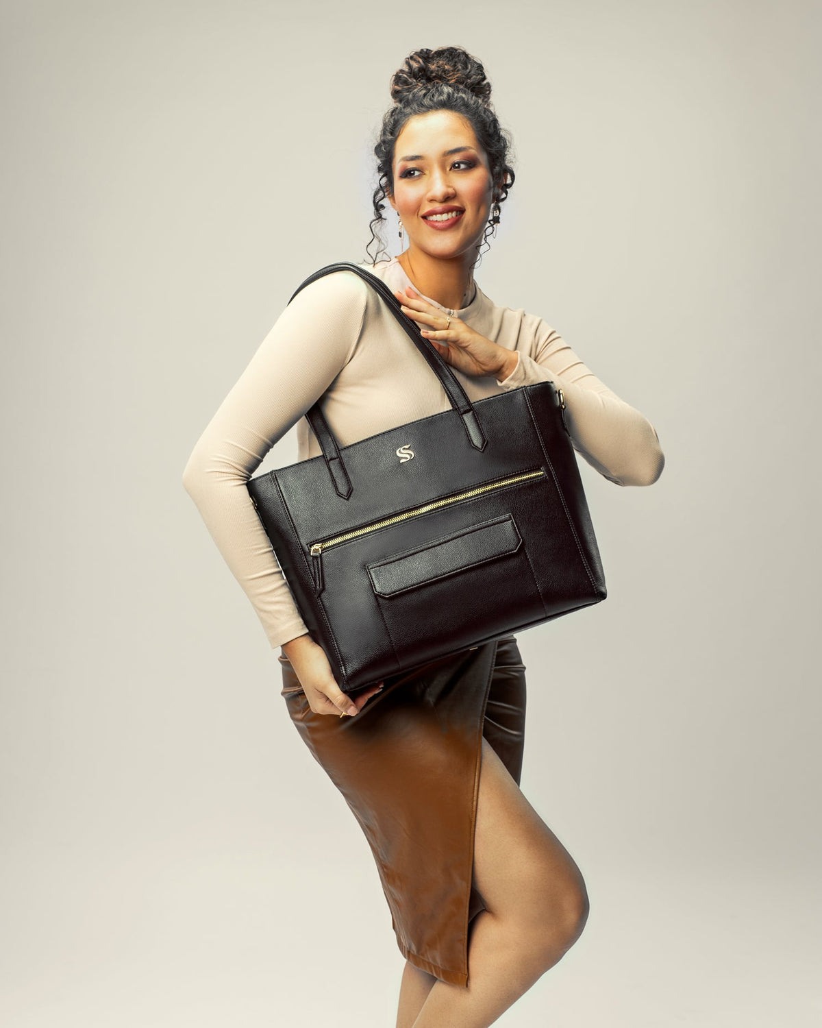 Black tote bag worn by model with top handles and crossbody strap, gold zipper accents