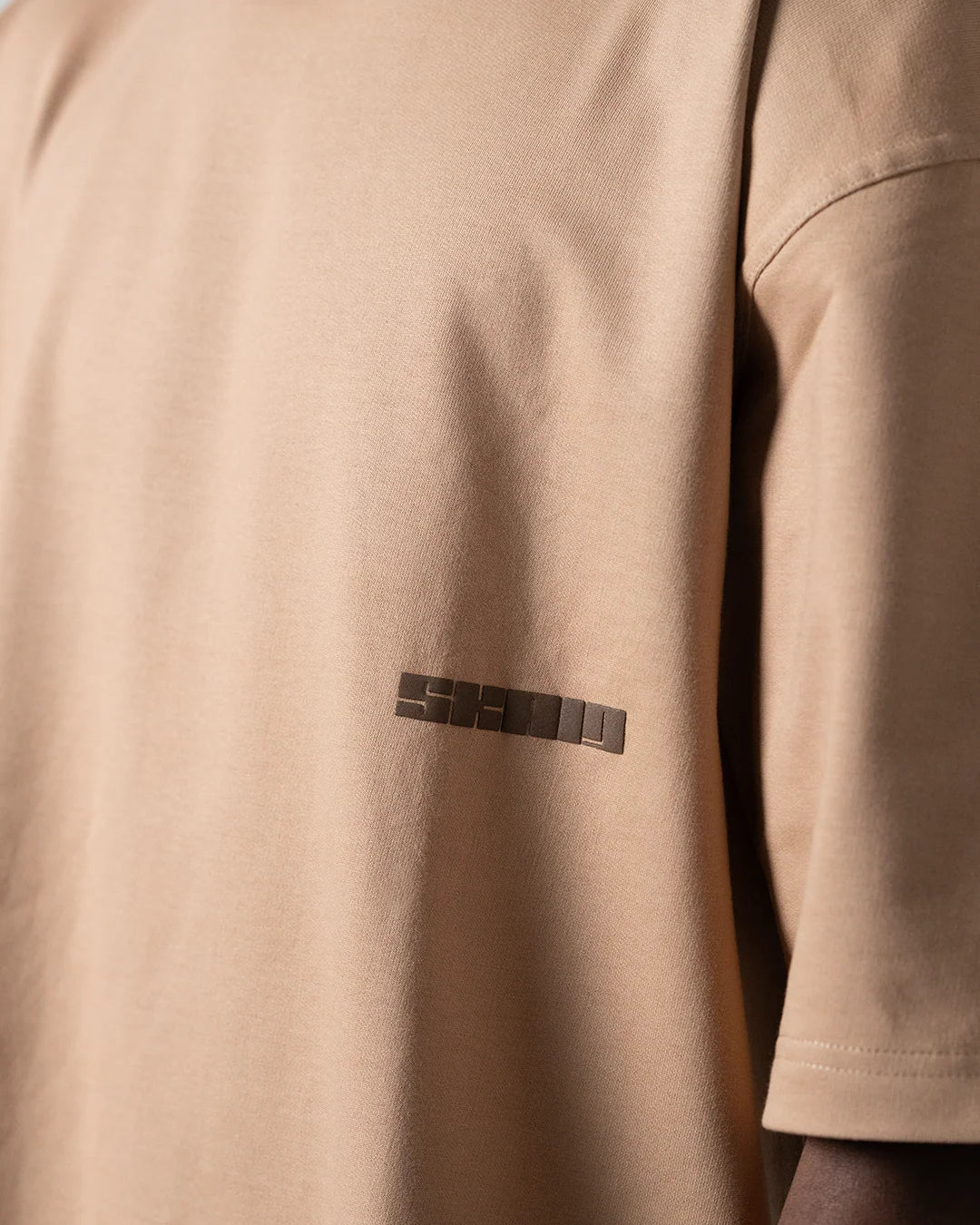 Beige T-shirt chest logo patch detail in dark print.