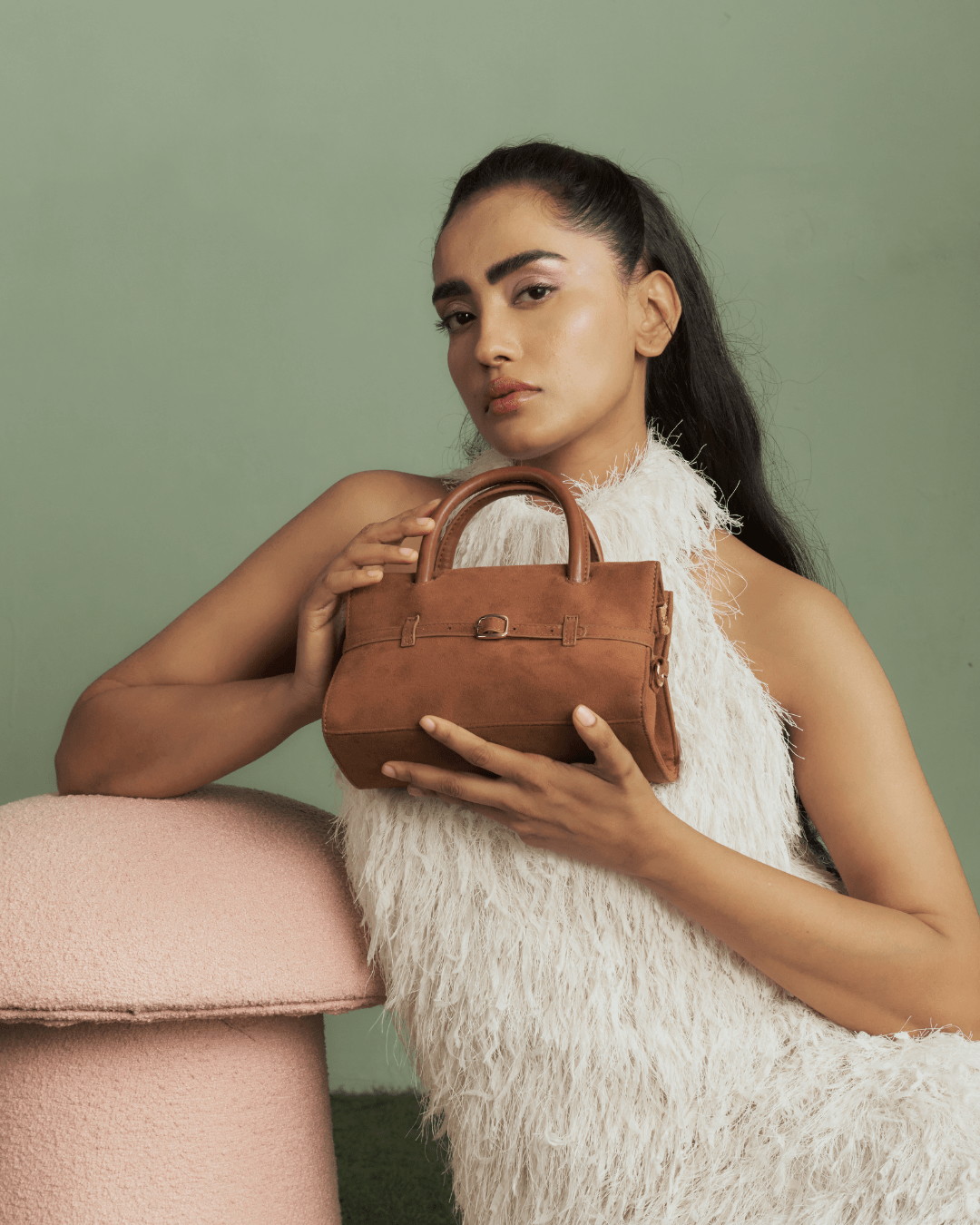 Tan mini handbag held by model, with top handles and front buckle strap.
