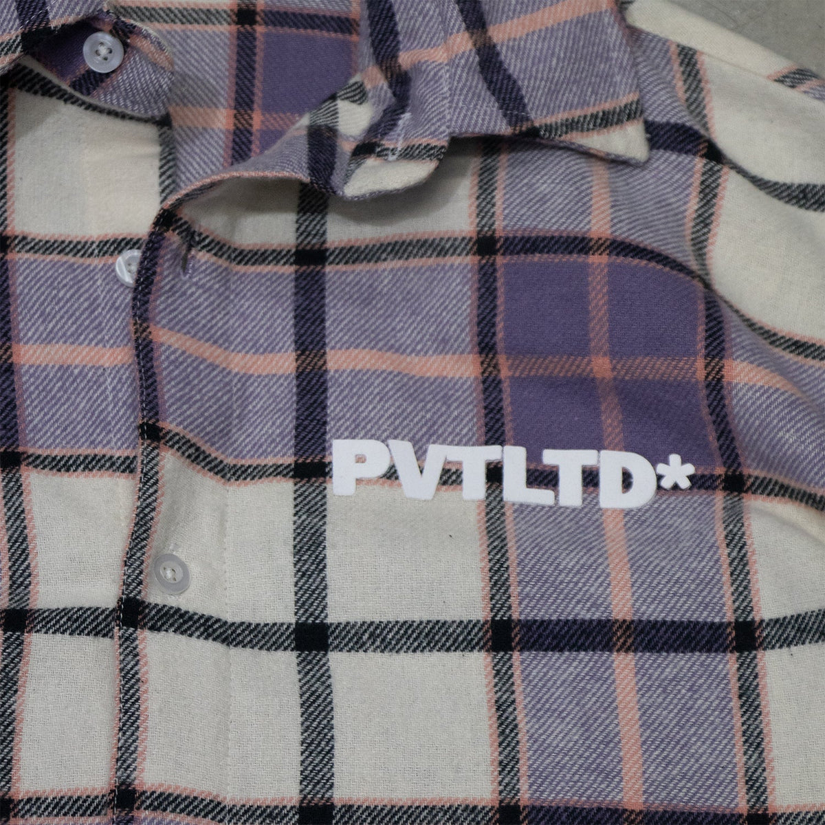 Close-up of PVT LTD* logo on purple and white plaid button-up shirt
