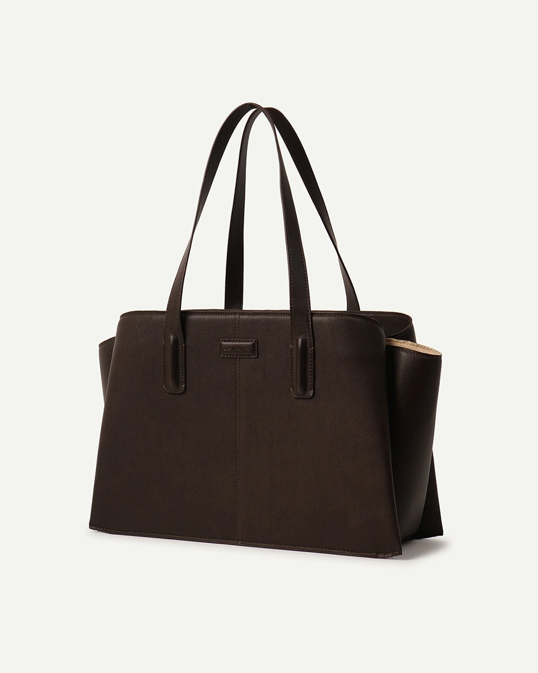 Dark brown leather tote bag with top handles and front logo patch, side view