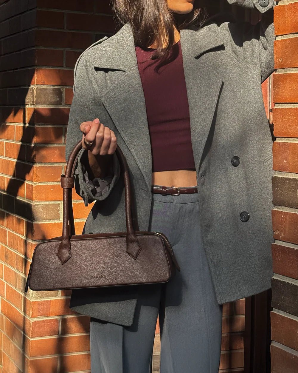Dark brown handbag with two top handles, held in hand at waist