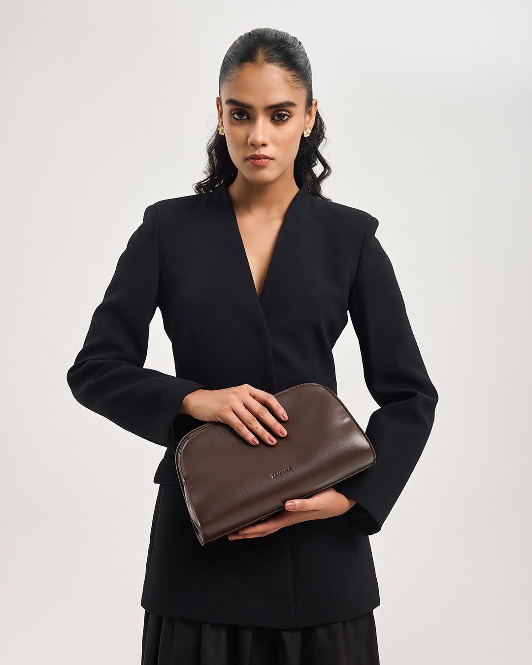 Dark brown leather clutch bag with short strap, carried by model in black blazer