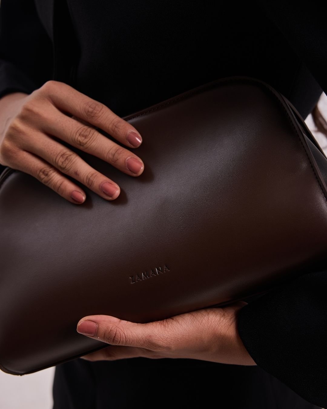 Dark brown leather clutch with smooth finish and small embossed logo, held with hands