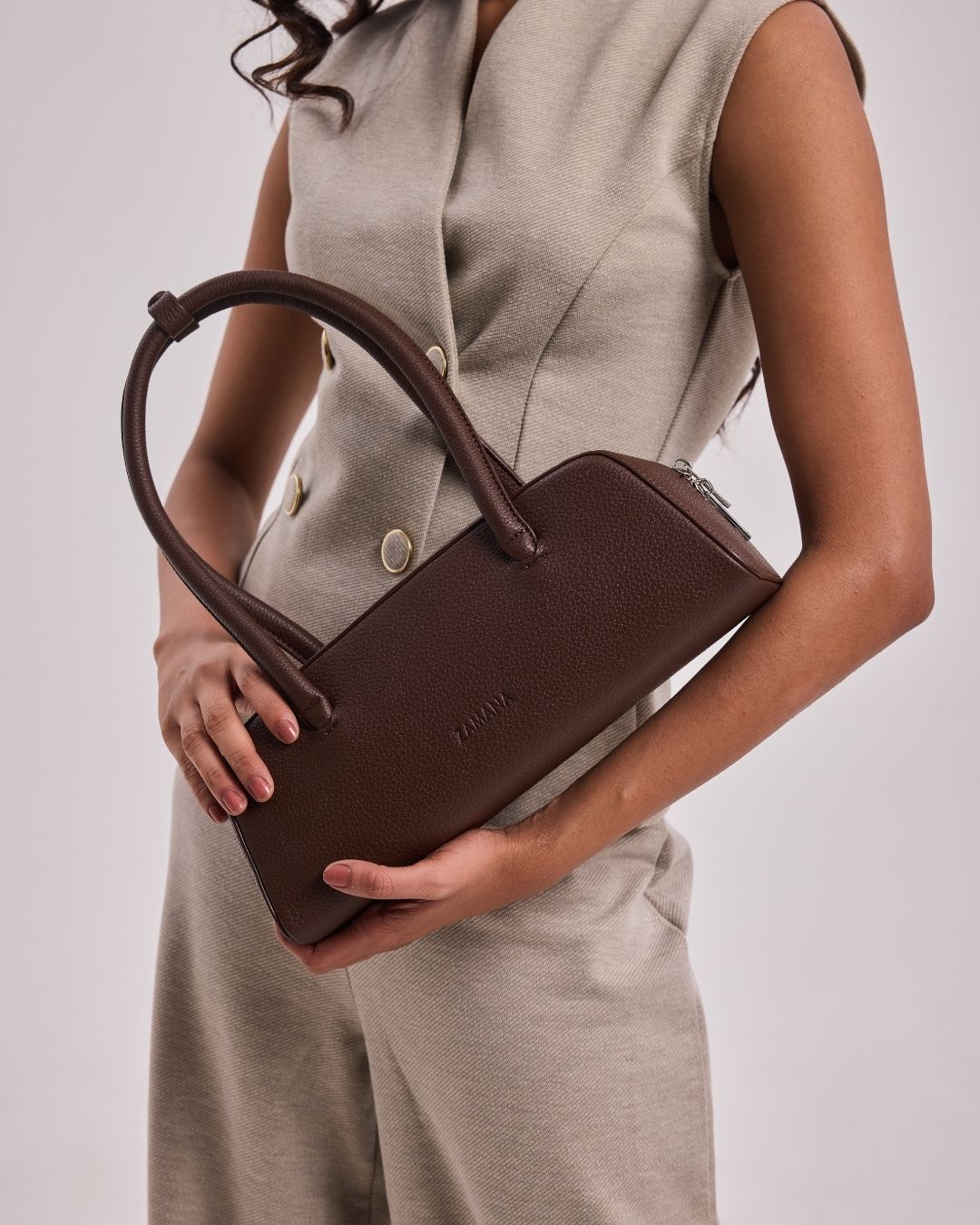 Dark brown top-handle handbag held in hand, with metal zipper and stitched logo detail.